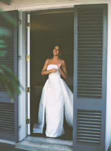Smiling woman in a strapless white gown stands in a doorway with gray shutters, lit by soft light before her Louisiana wedding.