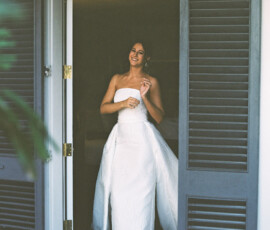 Smiling woman in a strapless white gown stands in a doorway with gray shutters, lit by soft light before her Louisiana wedding.