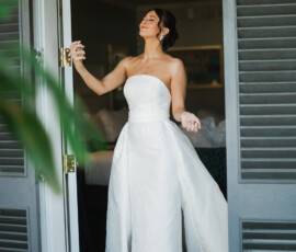 Smiling woman in a strapless white gown stands in a doorway, eyes closed, with greenery in the foreground.