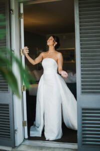 Smiling woman in a strapless white gown stands in a doorway, eyes closed, with greenery in the foreground.