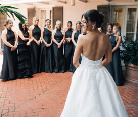 Bride in white strapless gown stands before smiling bridesmaids in black dresses at an outdoor Gulf South wedding.