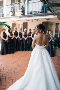 Bride in white strapless gown stands before smiling bridesmaids in black dresses at an outdoor Gulf South wedding.