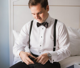 Man in bow tie and suspenders sits on bed, looking down and reflecting before Gulf South wedding ceremony.