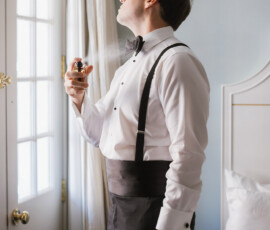 Man in formalwear sprays cologne by a window in a softly lit bedroom before his Louisiana wedding.