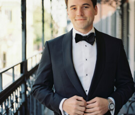 Man in black tuxedo with bow tie smiles on balcony, sunlit street and railings hint at a lively New Orleans wedding.