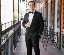 Man in black tuxedo poses confidently on a brick balcony with railings and chairs, ideal Gulf South wedding style.