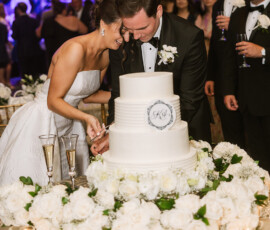 Bride and groom laughing as they cut their white wedding cake, with guests and white roses at a Louisiana wedding.