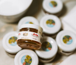 Small Maria Honey jar stacked atop others with white lids and colorful labels on a light table—great for wedding inspiration.