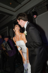 Bride and groom laughing on dance floor in Louisiana, both in novelty necklaces; saxophonist band plays behind them.