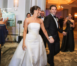 Bride in white strapless gown and groom in black tux walk into elegant New Orleans ballroom as guests applaud.
