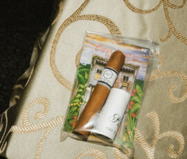 A cigar and container in a clear bag with house artwork rest on elegant gold and cream fabric, evoking Louisiana wedding charm.