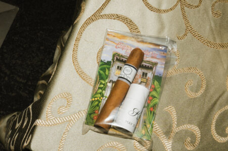 A cigar and container in a clear bag with house artwork rest on elegant gold and cream fabric, evoking Louisiana wedding charm.
