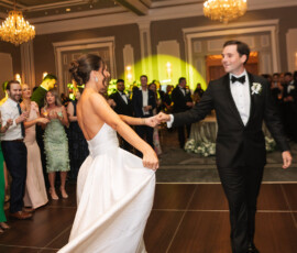 Bride and groom share a dance in a grand ballroom, her dress flowing as guests smile and chandeliers shine above.