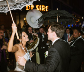 Bride and groom joyfully dance with decorated umbrellas at night, surrounded by musicians and a festive New Orleans crowd.