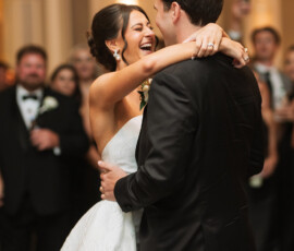 Bride and groom smiling and embracing on the dance floor at their New Orleans wedding, guests in formal attire nearby.