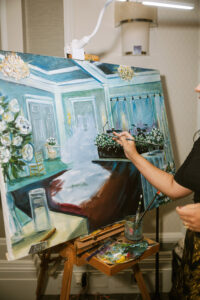 Artist painting Gulf South wedding scene with elegant decor, flowers, and tables; brush and art supplies visible.