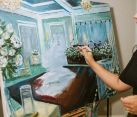 Artist painting Gulf South wedding scene with elegant decor, flowers, and tables; brush and art supplies visible.