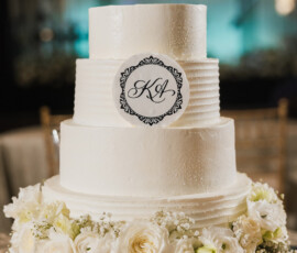Elegant three-tiered white wedding cake with monogram K A topper, silver stand, white roses, and baby’s breath accents.