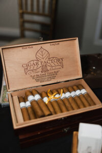 Open cigar box labeled “Cigar Factory New Orleans” with cigars, one tied by yellow ribbon; chair and dark surface in background.