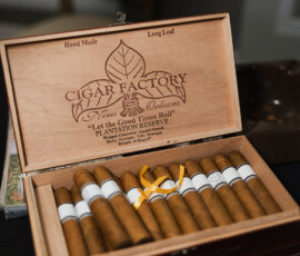 Open cigar box labeled “Cigar Factory New Orleans” with cigars, one tied by yellow ribbon; chair and dark surface in background.