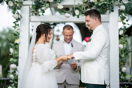 Bride and groom exchange rings under a flower arch, smiling, as an officiant conducts their New Orleans wedding ceremony.