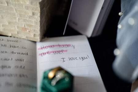 Notebook with “How lucky are we?” crossed out and “I love you!” written below, beside a green ring box, pearls, and fabric.