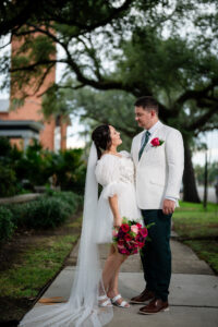 Bride in short white dress and veil smiles at groom; outdoors on a green path with pink bouquet, wedding inspiration.