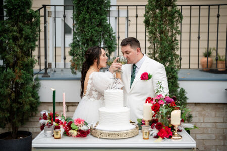 Bride and groom in white sip wine by a table with white cake, red flowers, candles, and greenery at their Gulf South wedding.
