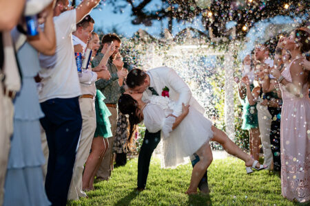 A groom dips and kisses his bride in a short white dress as guests blow bubbles at their outdoor New Orleans wedding.