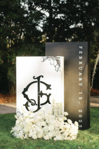 Custom monogram wedding sign with white floral arrangement displayed outdoors at the reception entrance.