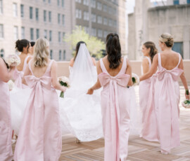 Bride in white gown with bridesmaids in pale pink dresses and bows on a city rooftop, perfect for Gulf South wedding ideas.