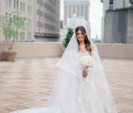 Bride in white lace gown with long veil and bouquet on a city rooftop, celebrating an elegant New Orleans wedding.