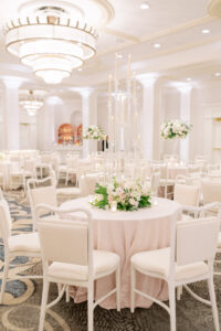 Round reception table featuring tall glass candlesticks, soft blush florals, and white chairs inside a grand ballroom.