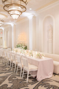 Long banquet table styled with blush linens, garden florals, and candlelight beneath ornate chandeliers.