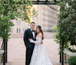 Bride in off-shoulder lace gown and groom in black tux on rooftop under floral arch at elegant Louisiana wedding with city backdrop.