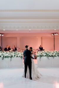 Bride and groom sharing their first dance on a white dance floor as a live band performs behind lush white florals.