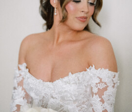 Bride in off-shoulder white lace dress holding pale bouquet, looking down with soft makeup and styled hair at New Orleans wedding.