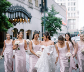 Bride in white gown and bridesmaids in purple dresses with bouquets walk a city street before their Louisiana wedding ceremony.
