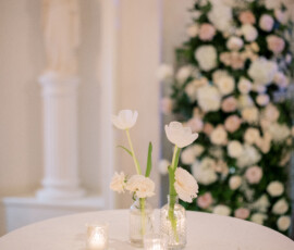 Elegant round table with white cloth, vases of white flowers, lit candles, floral arrangement, and statue in background.