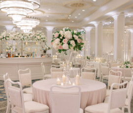 Elegant ballroom wedding reception with pink tablecloth, white chairs, floral centerpiece, candles, and chandeliers.