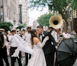 Bride and groom kiss in the street with feathered umbrellas, surrounded by a marching band at a vibrant Louisiana wedding.