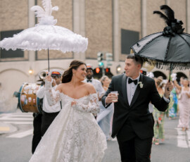 Bride and groom with feathered umbrellas lead musicians in a joyful New Orleans wedding street parade.