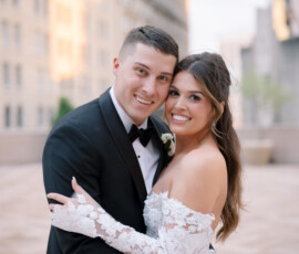 Bride and groom in New Orleans embrace and smile outdoors on their wedding day with city buildings in the background.