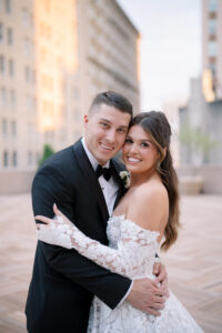Bride and groom embracing on an outdoor terrace, smiling at the camera in classic wedding attire with the city skyline softly blurred behind them.