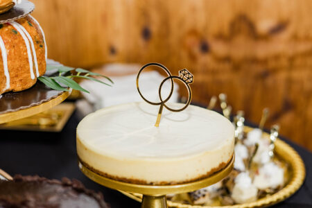 Cheesecake on a gold stand with a diamond ring topper, surrounded by desserts and a wooden background—ideal for Louisiana weddings.