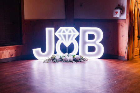 Illuminated J♡B letters with a diamond ring O and white flowers—romantic Gulf South wedding décor on wood floors.