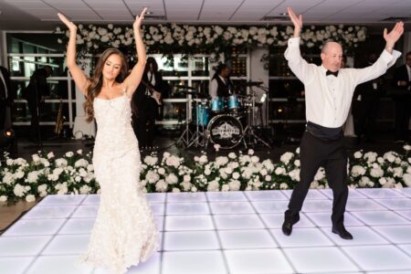 Bride and groom dance with raised arms on a lit floor at an elegant Louisiana wedding with band and floral decor.