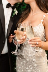 Bride and groom in formal attire clink champagne glasses, her engagement ring visible—wedding inspiration from Louisiana.