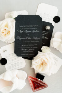 Black wedding invitation suite with white script, white roses, rings, wax seals, shoes, and red Prada perfume on light background.