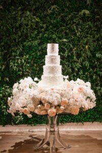 Five-tier white wedding cake on glass table, surrounded by soft flowers and greenery, at a Louisiana ceremony.
